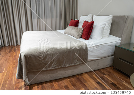 Modern hotel bedroom with a neatly made double bed, white linens, beige upholstered frame and red accent pillows on wooden floor 135459740