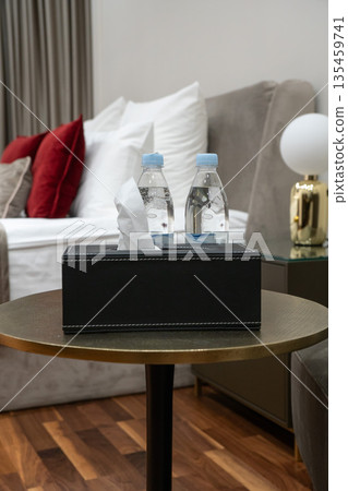 Hotel bedside table with bottled water and tissue box in a modern guest room, featuring a comfortable bed, decorative pillows and soft lighting 135459741