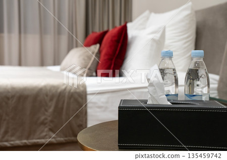 Hotel bedside table with bottled water and tissue box in a modern guest room, featuring a comfortable bed, decorative pillows and soft lighting 135459742