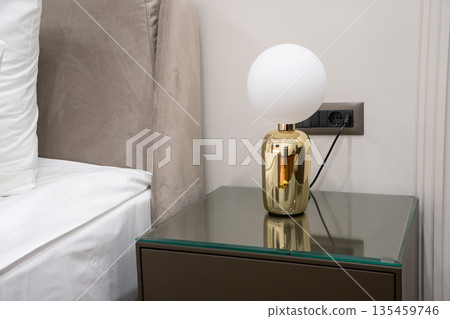 Modern bedside table with glass top and gold table lamp featuring a white spherical shade in a contemporary hotel bedroom interior Modern bedside table with glass top and gold table lamp featuring a white spherical shade in a contemporary hotel bedroom interior 135459746