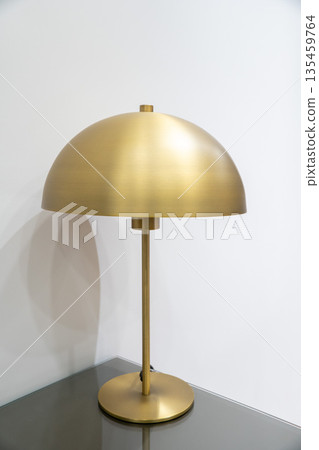 Modern gold metal table lamp with dome-shaped shade standing on a bedside table against a white wall. Suitable for concepts of interior design 135459764