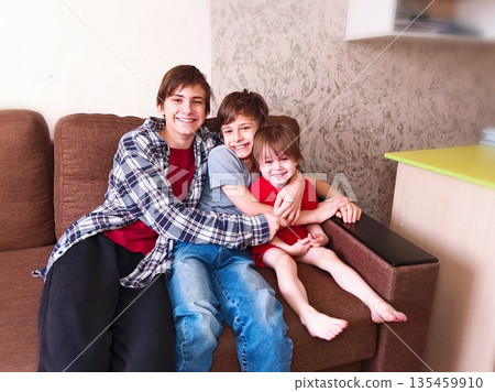 Three Brothers Hugging on Couch Smiling. Happy teenage and young brothers cuddle together on cozy sofa, big smiles, warm family moment indoors. 135459910