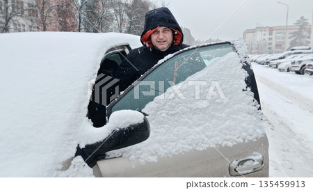 Smiling Man in Snowy Car Window. Cheerful man opens car door, smiles from inside snow-covered vehicle during winter blizzard in urban area. 135459913
