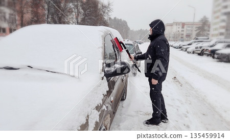 Man Cleaning Snow from Car Winter. Man in black jacket brushes thick snow off car window with red scraper during heavy snowfall in city parking. Man Cleaning Snow from Car Winter. Man in black jacket brushes thick snow off car window with red scraper during heavy snowfall in city parking. 135459914