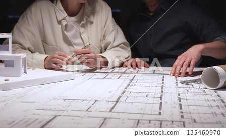 Architects and engineers collaborating on a new building project, planning structural details and design concepts using blueprints and a scaled architectural model on a desktop Architects and engineers collaborating on a new building project, planning structural details and design concepts using blueprints and a scaled architectural model on a desktop 135460970