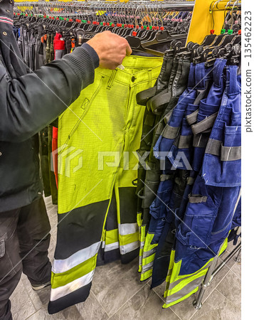 A customer in a workwear store browses the selection, holding up a pair of high-visibility yellow safety trousers to inspect them before buying. High quality photo 135462223