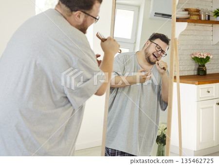 Overweight happy man singing with comb as microphone before mirror at home Overweight happy man singing with comb as microphone before mirror at home 135462725