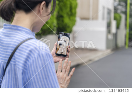 畫面中，一位中年婦女外出時，正透過智慧型手機的寵物攝影機查看她的寵物狗的狀況。 135463425