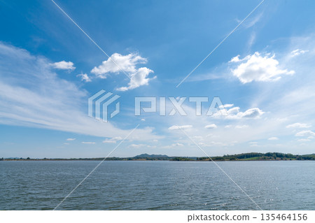 Wide landscape of a calm blue lake under a bright sunny sky with white wispy clouds. Scenic view of a reservoir surrounded by distant green hills and trees. Peaceful natural outdoor background. Wide landscape of a calm blue lake under a bright sunny sky with white wispy clouds. Scenic view of a reservoir surrounded by distant green hills and trees. Peaceful natural outdoor background. 135464156