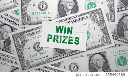 A white card with the message WIN PRIZES in green text sits atop a pile of US one-dollar bills, representing concepts of luck, winning, rewards, and financial incentives 135464389
