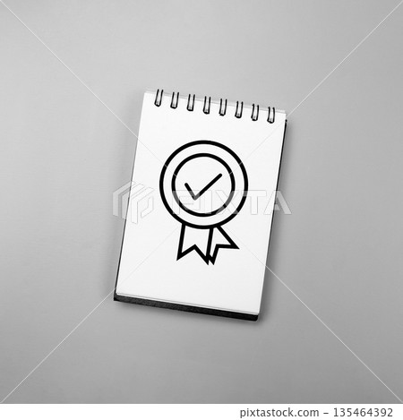High angle shot of a white spiral notebook featuring a black line art quality seal icon with a check mark, set against a solid grey background. Perfect for business and quality control concepts High angle shot of a white spiral notebook featuring a black line art quality seal icon with a check mark, set against a solid grey background. Perfect for business and quality control concepts 135464392