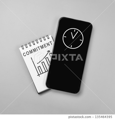 Top view flat lay of a notebook with 'COMMITMENT' and a growth graph under a smartphone displaying a clock icon, representing time management and business success Top view flat lay of a notebook with 'COMMITMENT' and a growth graph under a smartphone displaying a clock icon, representing time management and business success 135464395