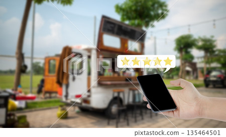Close-up of a hand holding a smartphone with a five-star rating overlay against a blurred food truck background, representing customer feedback and excellent service reviews Close-up of a hand holding a smartphone with a five-star rating overlay against a blurred food truck background, representing customer feedback and excellent service reviews 135464501