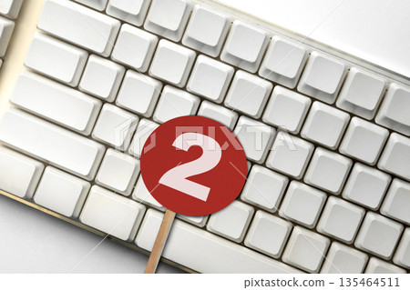A close-up view of a white computer keyboard with a red circular sign featuring the number two placed on top, representing steps, rankings, or notifications A close-up view of a white computer keyboard with a red circular sign featuring the number two placed on top, representing steps, rankings, or notifications 135464511