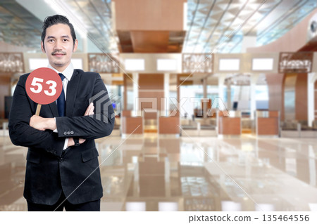 Indonesian southeast asian businessman in a formal suit holds a red circular sign with the number 53, standing in a blurred modern airport terminal or corporate office lobby 135464556