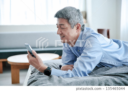 Senior man lying on the bed and looking at his smartphone 135465044