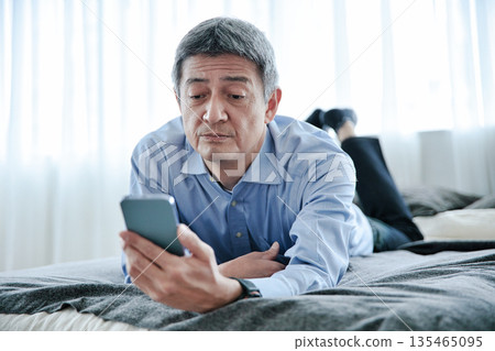 Senior man lying on the bed and looking at his smartphone Senior man lying on the bed and looking at his smartphone 135465095