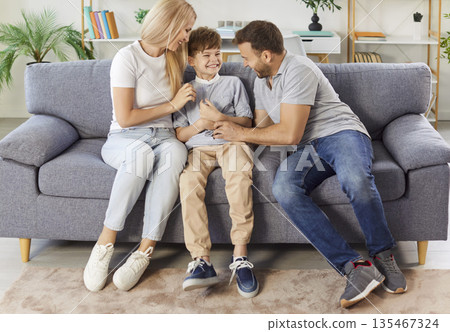 Happy family at home, cheerful excited parents tickling beloved kid son, playing with child on sofa Happy family at home, cheerful excited parents tickling beloved kid son, playing with child on sofa 135467324