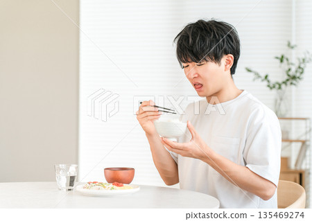 一位年輕的亞洲男子在家裡的餐廳吃著難以下嚥的白米飯（進口米、陳米、米短缺）。 135469274