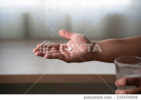 man hand holds a white pill or capsule and a glass of water at home, eating vitamin on time, medicine daily vitamins ginseng or calcium support health, wellness, health and care. 135470998