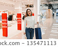 Happy Woman with Shopping Bags Stands Confidently in Store Aisle 135471113