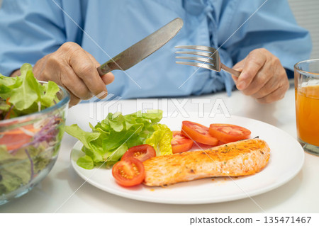 Asian elderly woman patient eating salmon stake and vegetable salad for healthy food in hospital. 135471467