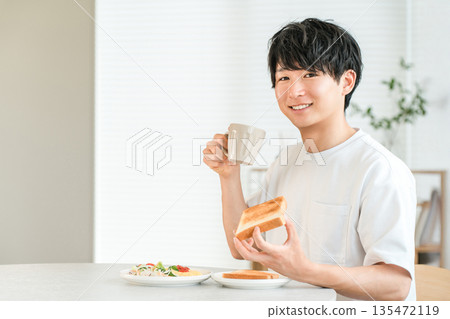 一位年輕的亞洲男子一邊喝咖啡，一邊吃著吐司麵包和沙拉當早餐。 135472119