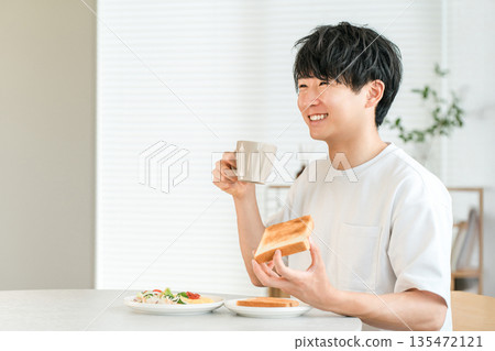 一位年輕的亞洲男子一邊喝咖啡，一邊吃著吐司麵包和沙拉當早餐。 135472121
