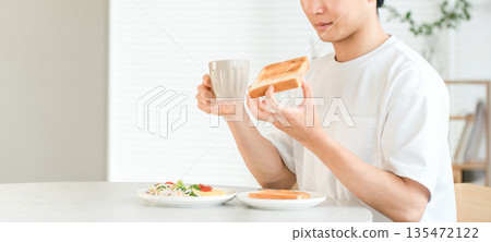一位年輕的亞洲男子一邊喝咖啡,一邊吃著吐司麵包和沙拉當早餐。 一位年輕的亞洲男子一邊喝咖啡,一邊吃著吐司麵包和沙拉當早餐。 135472122