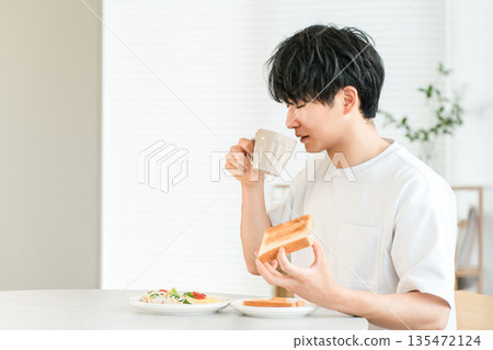 一位年輕的亞洲男子一邊喝咖啡，一邊吃著吐司麵包和沙拉當早餐。 135472124