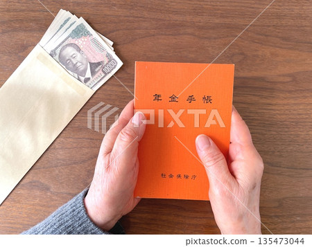 Elderly woman's hands holding a pension book and banknotes on a wooden table Elderly woman's hands holding a pension book and banknotes on a wooden table 135473044