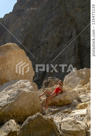Beach woman rocks, young woman relaxes in red swimsuit on sun-drenched beach with massive cliffs. 135473139
