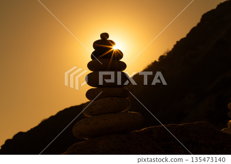 Cairn, sunset, balance. Stacked zen stones creating a silhouette with a dramatic sunburst on a rocky hillside. 135473140