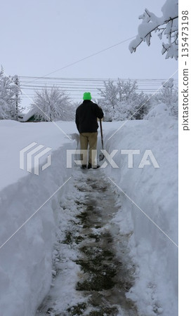 A vertical image of a man standing with his back to the ground with a shovel on a path made through large snowdrifts in a front yard in a rural area, clearing a path to the road after abnormal snowfal A vertical image of a man standing with his back to the ground with a shovel on a path made through large snowdrifts in a front yard in a rural area, clearing a path to the road after abnormal snowfal 135473198