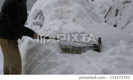 unrecognizable man uses a long-handled scraper to clear the front of a gray car, covered in deep snow after a severe storm. Clearing a car of heavy snow, starting with the front bumper and headlights 135473763