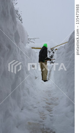 A man in dark clothing clears a path through high snowdrifts, creating a snow tunnel. The aftermath of an abnormal snowfall in a village. Manually clearing a path from heavy snow. 135473988