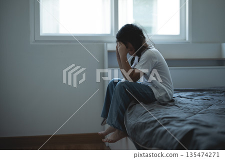 Young woman sitting on bed, feeling sad and distressed, covering face with hands, indoor setting, natural light coming through window, emotional moment captured Young woman sitting on bed, feeling sad and distressed, covering face with hands, indoor setting, natural light coming through window, emotional moment captured 135474271