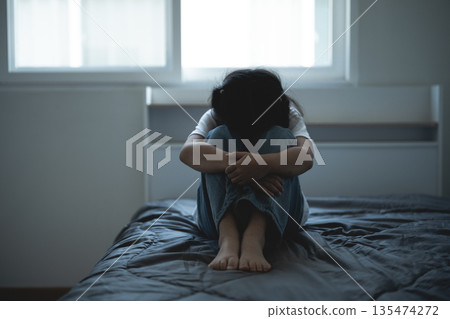 Emotional distress portrayed by a young girl sitting alone on a bed with her knees hugged close to her chest in a dimly lit room Emotional distress portrayed by a young girl sitting alone on a bed with her knees hugged close to her chest in a dimly lit room 135474272
