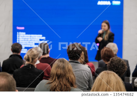 Presenter speaks to attentive audience during conference with large blue screen backdrop 135474682
