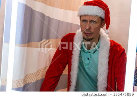 A middle-aged Caucasian man with short brown hair wears a red Santa hat and a red coat with white fur trim. He stands in a festive setting with soft lighting. A middle-aged Caucasian man with short brown hair wears a red Santa hat and a red coat with white fur trim. He stands in a festive setting with soft lighting. 135475099