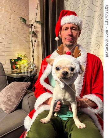 A man with light skin and short hair wears a Santa hat and red coat. He holds a small white Chihuahua dressed in reindeer antlers. The background is festive. A man with light skin and short hair wears a Santa hat and red coat. He holds a small white Chihuahua dressed in reindeer antlers. The background is festive. 135475108