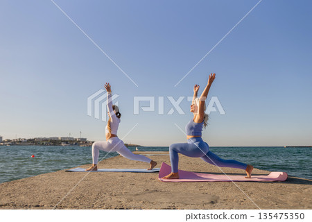 Yoga Women Beach Pose Fitness Outdoor - Two women practice yoga on a pier by the ocean. Yoga Women Beach Pose Fitness Outdoor - Two women practice yoga on a pier by the ocean. 135475350