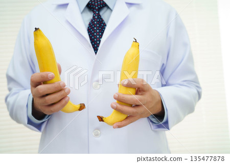 Male doctor stands holding a banana, penis health. Male doctor stands holding a banana, penis health. 135477878