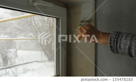 A hand with a brush begins to paint with white paint the plastered slope of a small rectangular window near a gray wall, with a view of the winter garden behind the glass A hand with a brush begins to paint with white paint the plastered slope of a small rectangular window near a gray wall, with a view of the winter garden behind the glass 135478183