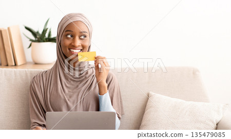 Full length portrait of black woman in hijab shopping on internet with credit card and laptop, indoors. Positive Muslim lady in traditional headscarf buying goods in online store Full length portrait of black woman in hijab shopping on internet with credit card and laptop, indoors. Positive Muslim lady in traditional headscarf buying goods in online store 135479085
