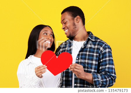 Valentines day, couple relationship, anniversary, love and romance. Smiling teen african american male and female looking at each other and hold big red paper heart, isolated on yellow background Valentines day, couple relationship, anniversary, love and romance. Smiling teen african american male and female looking at each other and hold big red paper heart, isolated on yellow background 135479173