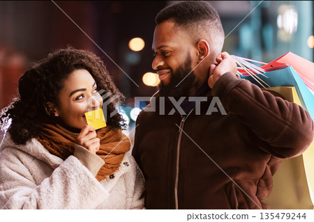 It's Shopping Time. Portrait of smiling black woman holding debit credit card near mouth and showing it to her husband, couple walking together in the evening and carrying bags, retail and purchase 135479244