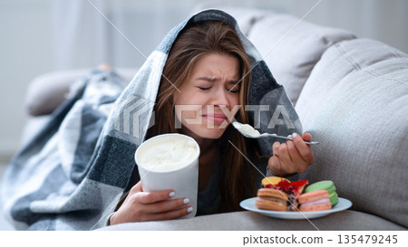 Upset young woman dealing with depression by eating ice cream and sweets under blanket on sofa. Stressed millennial lady coping with personal or relationship problem by comfort food Upset young woman dealing with depression by eating ice cream and sweets under blanket on sofa. Stressed millennial lady coping with personal or relationship problem by comfort food 135479245