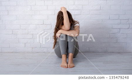 Desperate young lady crying from hopelessness on floor near white wall, blank space. Stressed millennial woman suffering from depression, PTSD or anxiety. Mental health care concept 135479248