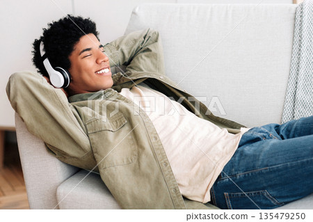 Happy black teen guy in headphones lying on comfy couch and listening to music at home, copy space. African American teenager spending lazy time on weekend, enjoying his favorite song 135479250